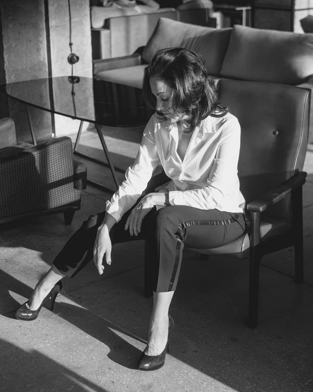 A stylish brunette woman sits in a chair in a monochrome setting, exuding elegance and confidence.