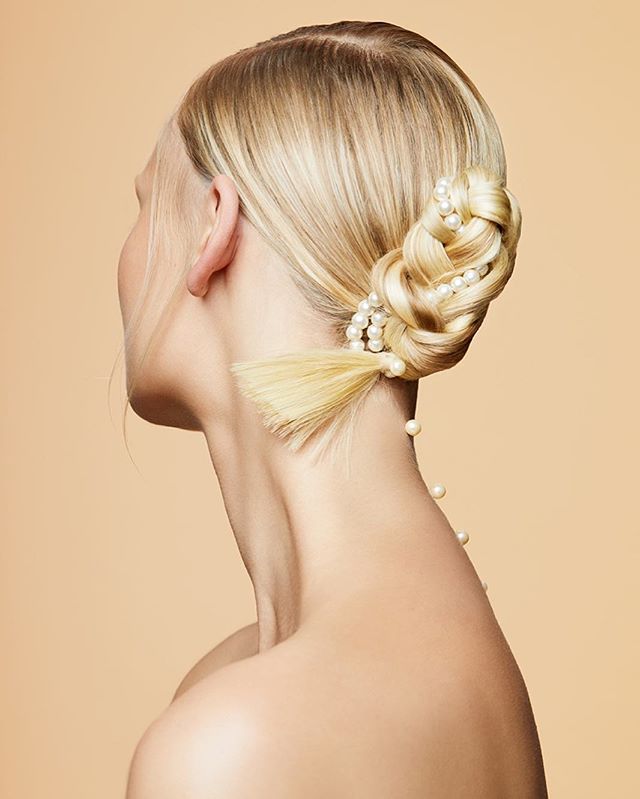 Elegant blonde woman with a braided hairstyle and pearls, photographed in a studio against a beige background.