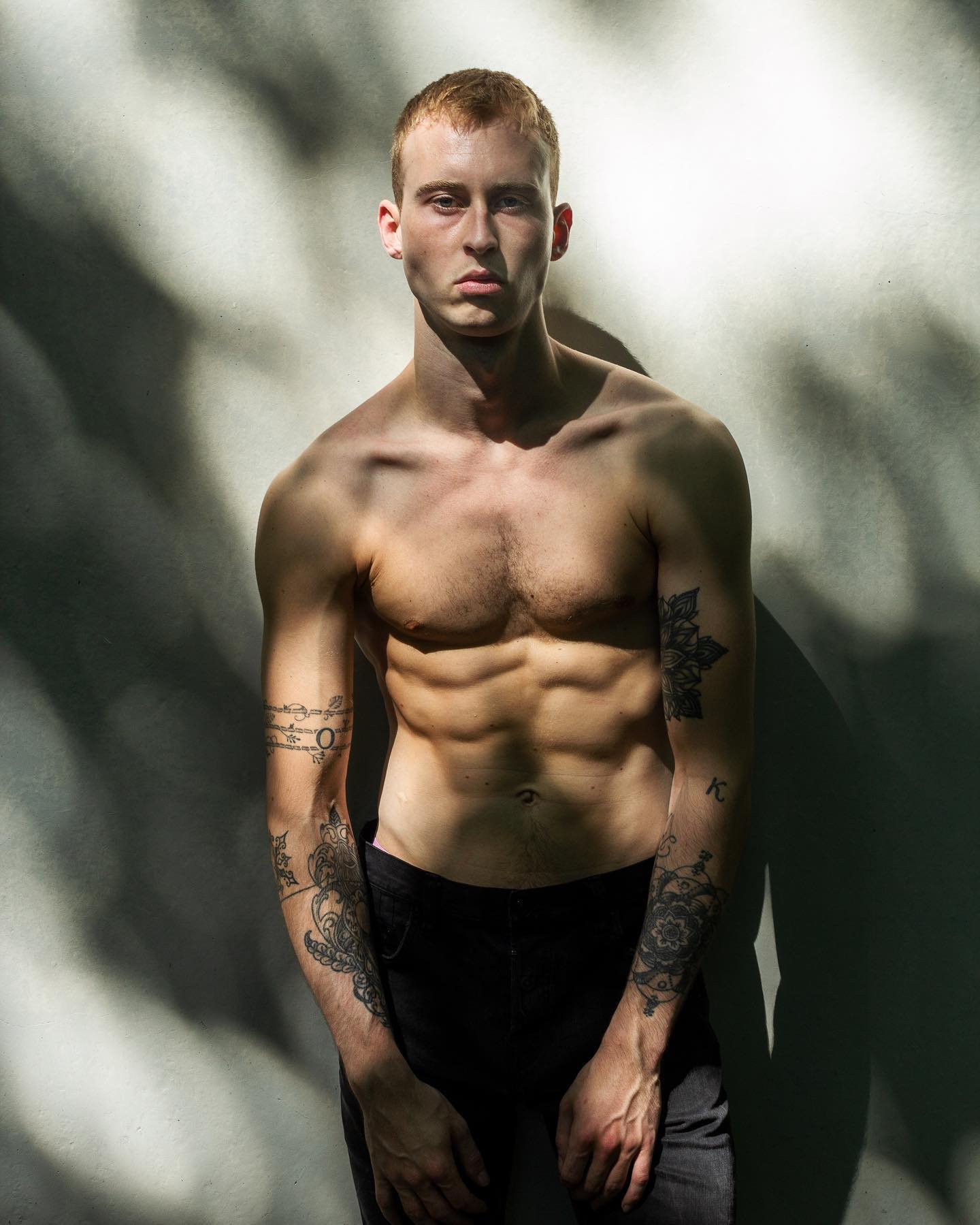 A muscular man with tattoos stands confidently in front of a gray wall bathed in natural light and shadows.