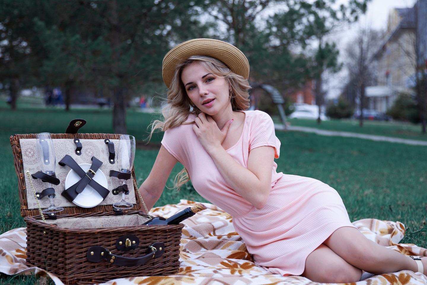 A blonde woman in a pink dress enjoys a relaxing picnic in a park with wine and a basket.