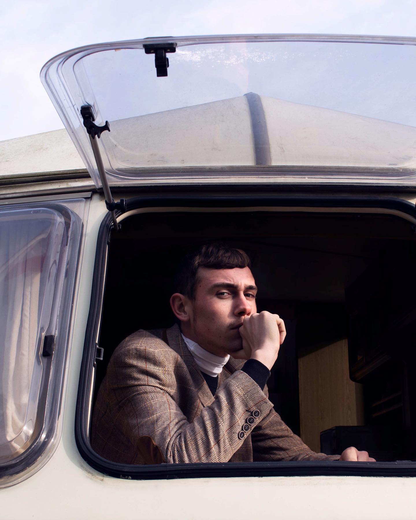 A contemplative man in a tweed jacket gazes out from the open window of a caravan.