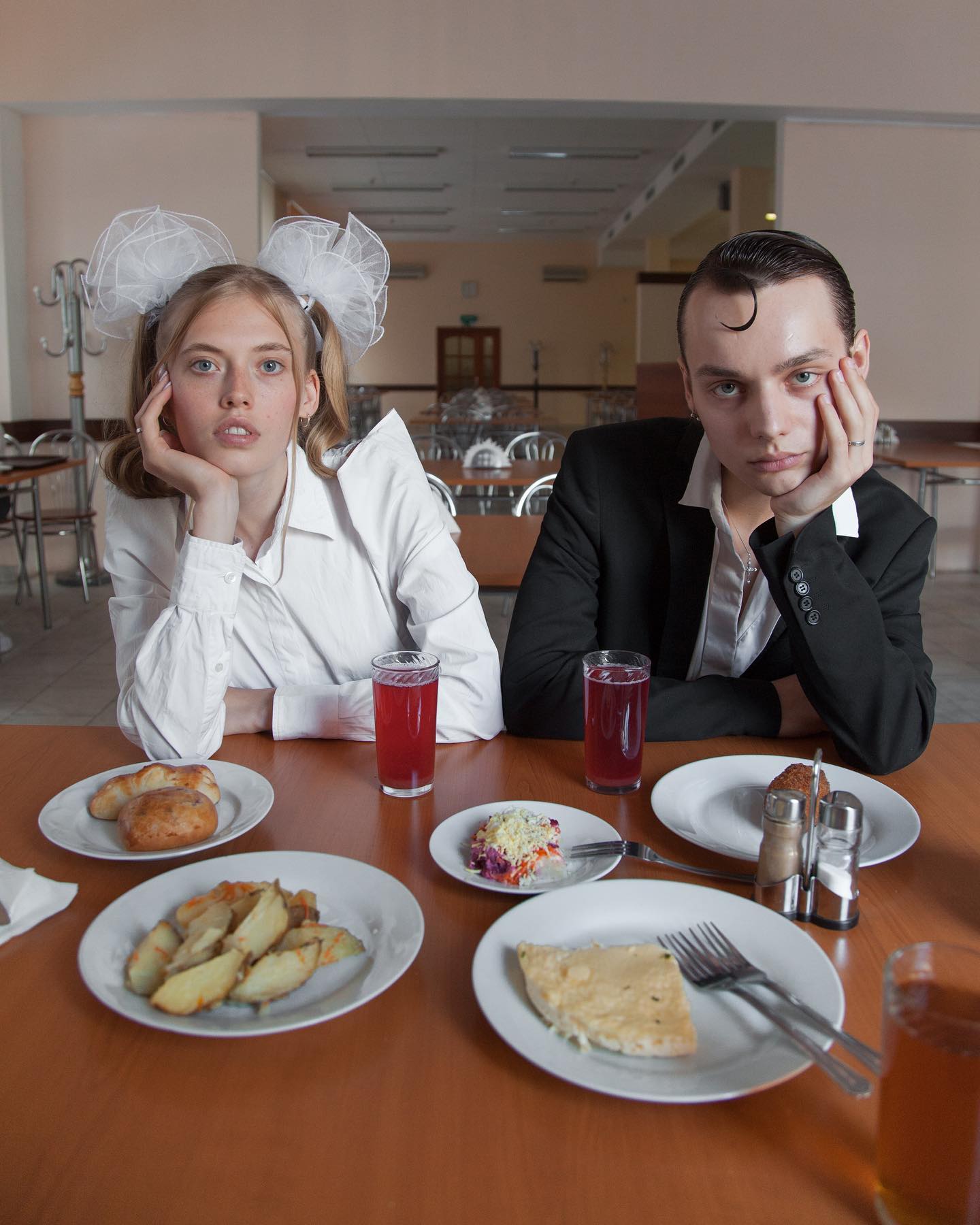A young couple sits at a cafeteria table with food and drinks, conveying a retro or vintage aesthetic.
