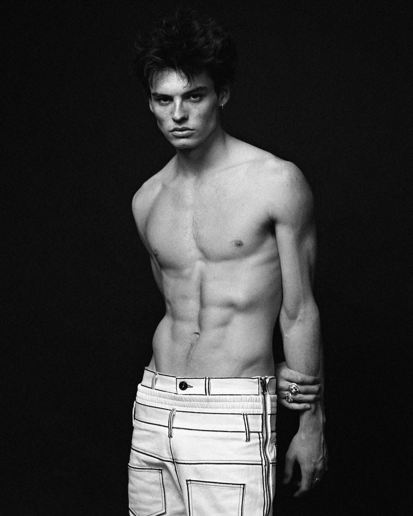 A shirtless man with defined abs poses in a black and white studio shot wearing white pants.