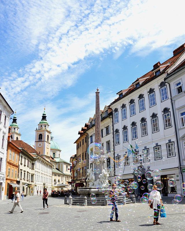 A sunny city square scene with people, buildings, a fountain, and many floating soap bubbles.