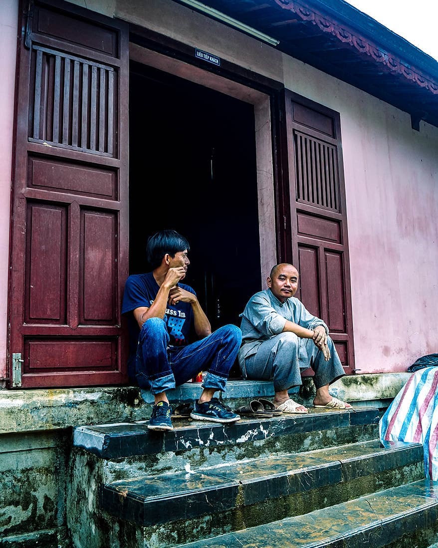 Two men sit on the steps of a building in an outdoor setting.