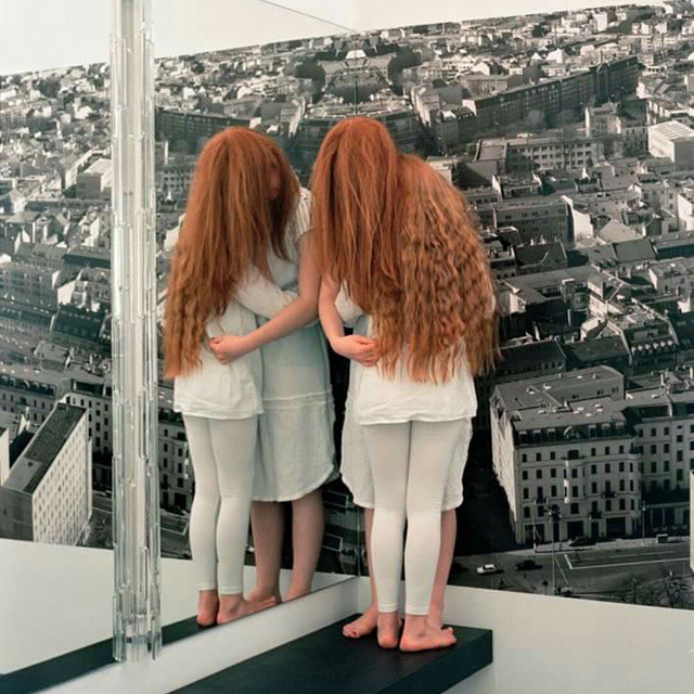 Two women with long red hair stand before a mirror, their reflections blending with a cityscape backdrop in an artistic scene.