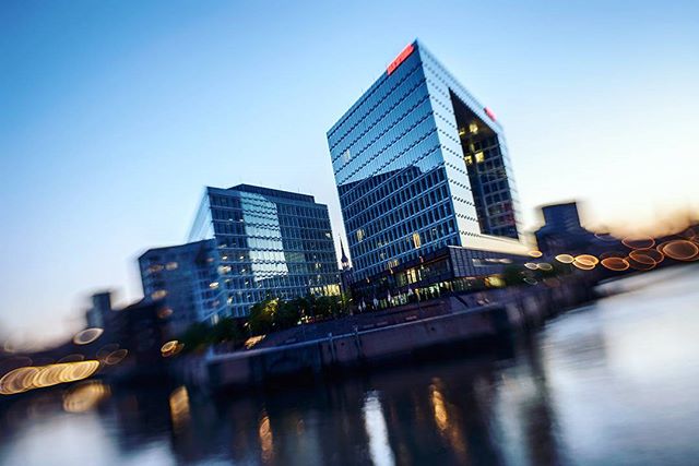 Modern office buildings reflected in the river create a sleek urban skyline, captured with a blurred, tilt-shift style.