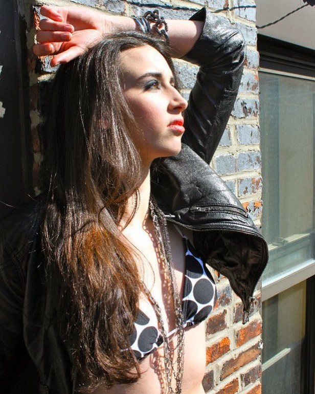 A confident woman in a bikini and leather jacket poses against a brick wall in sunlight.