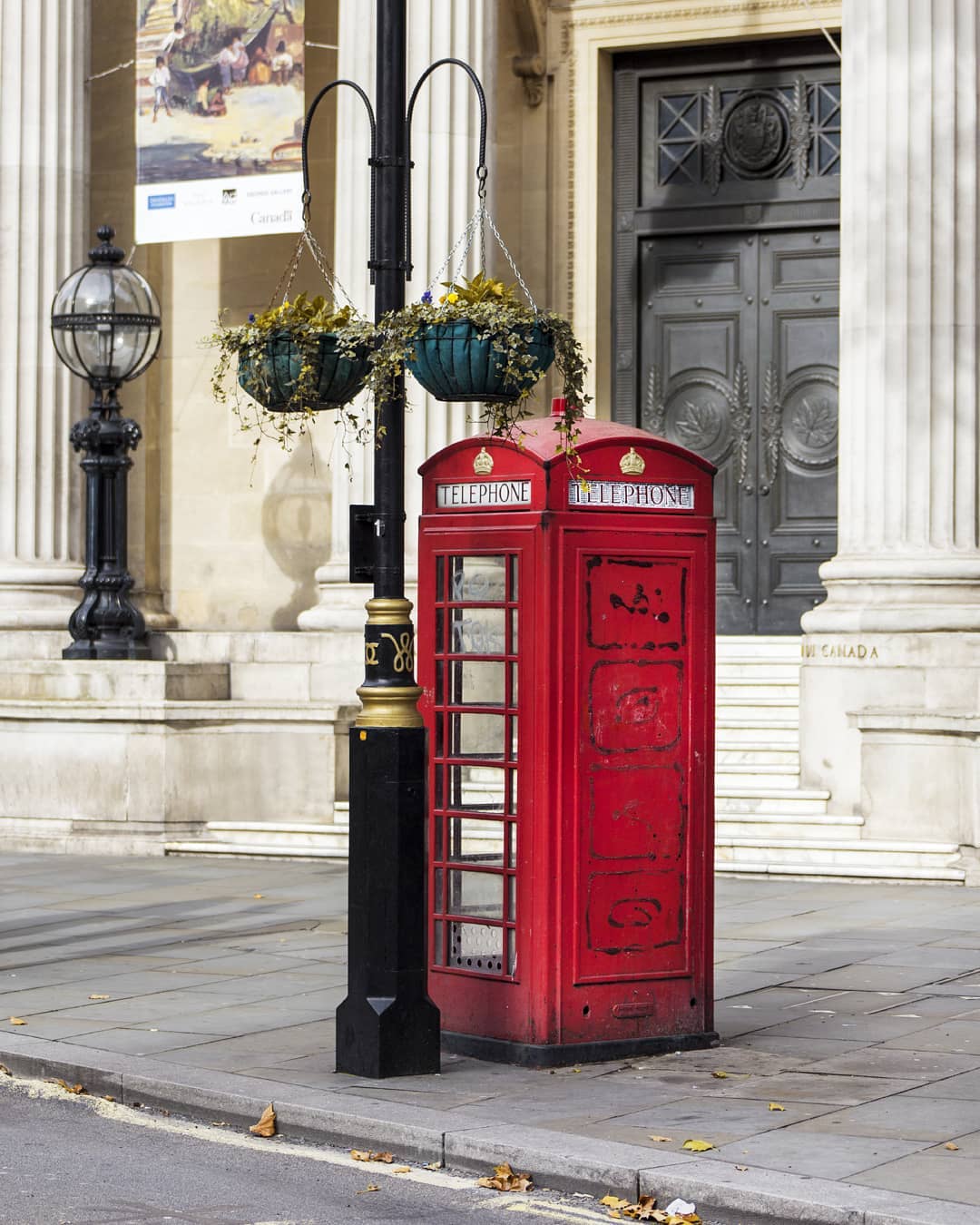 A classic red telephone booth stands on a city street next to a black lamp post with hanging flower baskets.