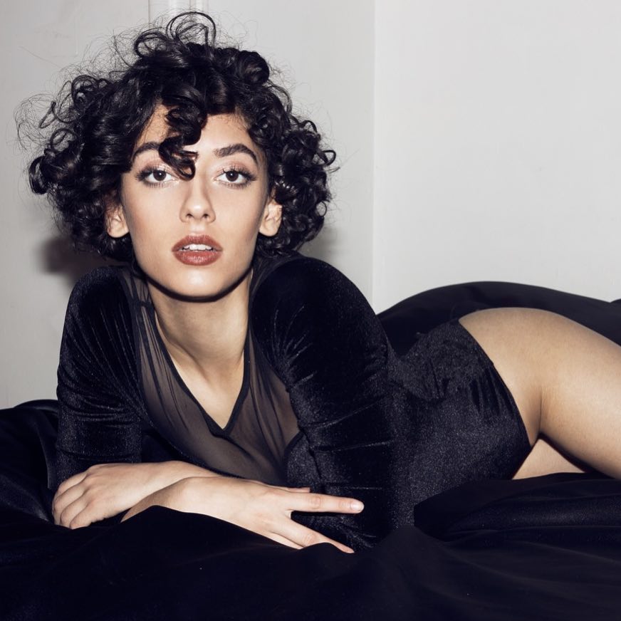 A brunette woman with curly hair lies on a black satin sheet in a black velvet and sheer bodysuit.