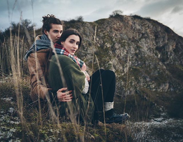 A couple embrace in a natural setting. Wearing scarves and outdoor clothes, with a rocky hill in the background.
