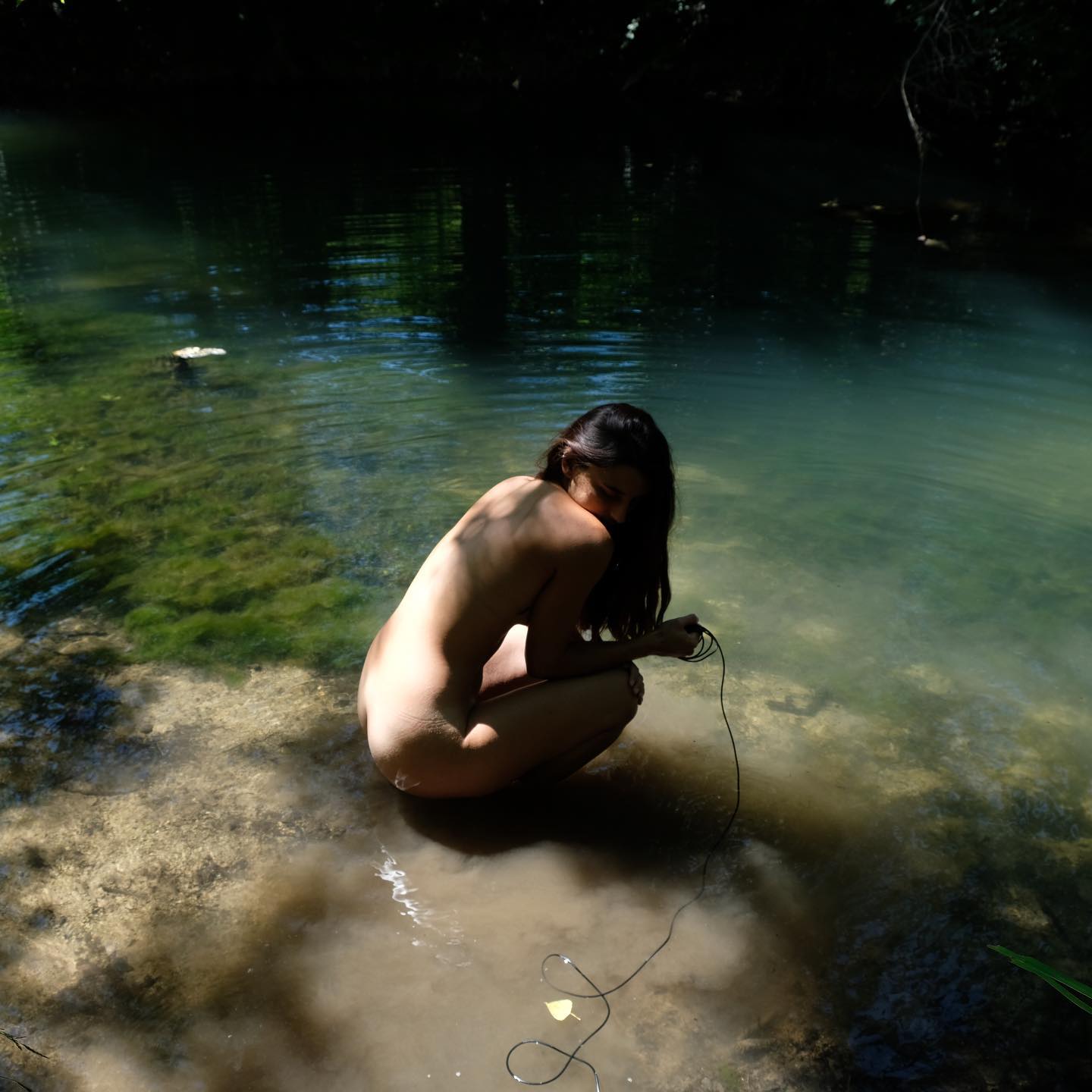 A nude woman sits serenely by a river in a lush forest, bathing in nature's beauty.