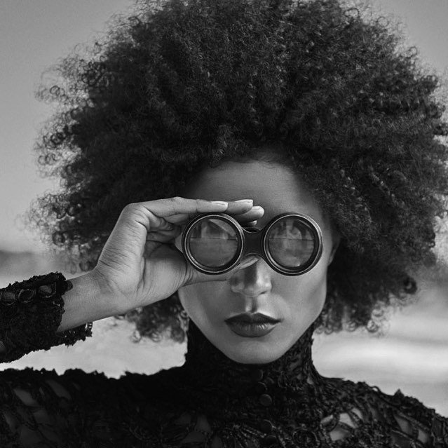 A stylish woman with afro hair gazes through binoculars in a black and white editorial shot.