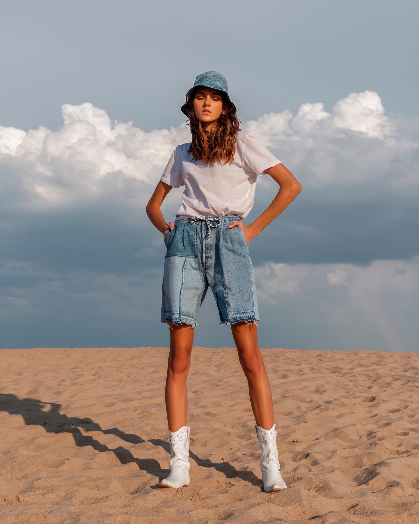 A woman in denim shorts, a white t-shirt, and white boots stands on a sand dune with a cloudy sky behind her.
