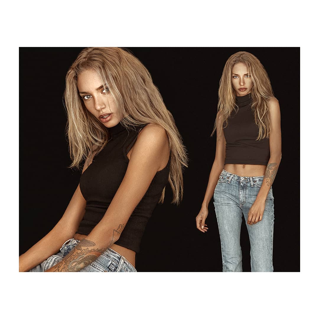 A blonde woman in a black top and jeans poses in a studio with a black background.