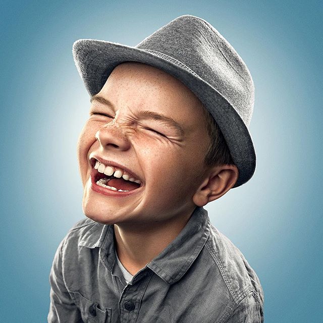 A young boy in a gray hat and shirt is laughing with his eyes closed against a blue background.