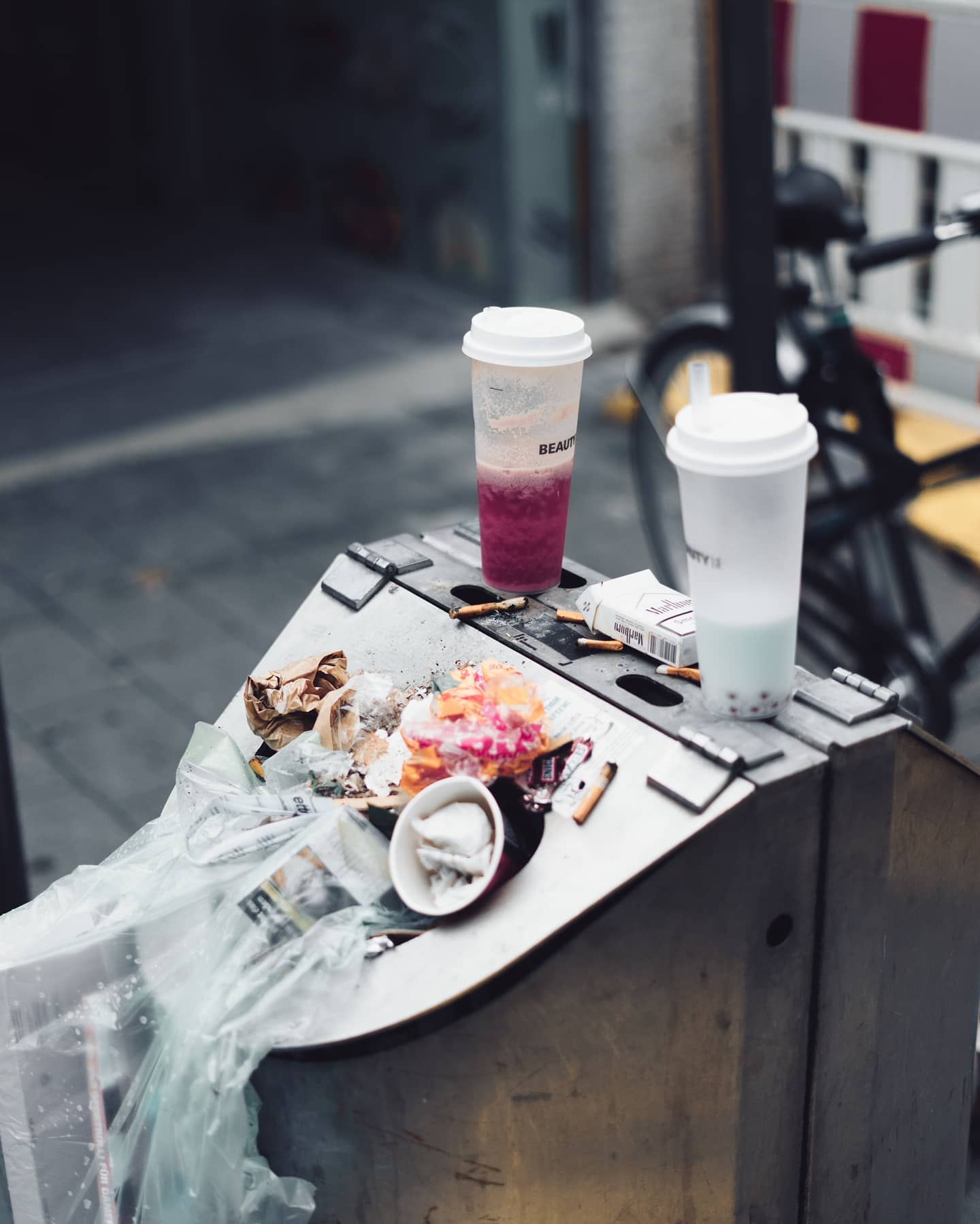 A trash can overflows with litter, cigarette butts, and discarded drinks, highlighting urban waste and pollution.