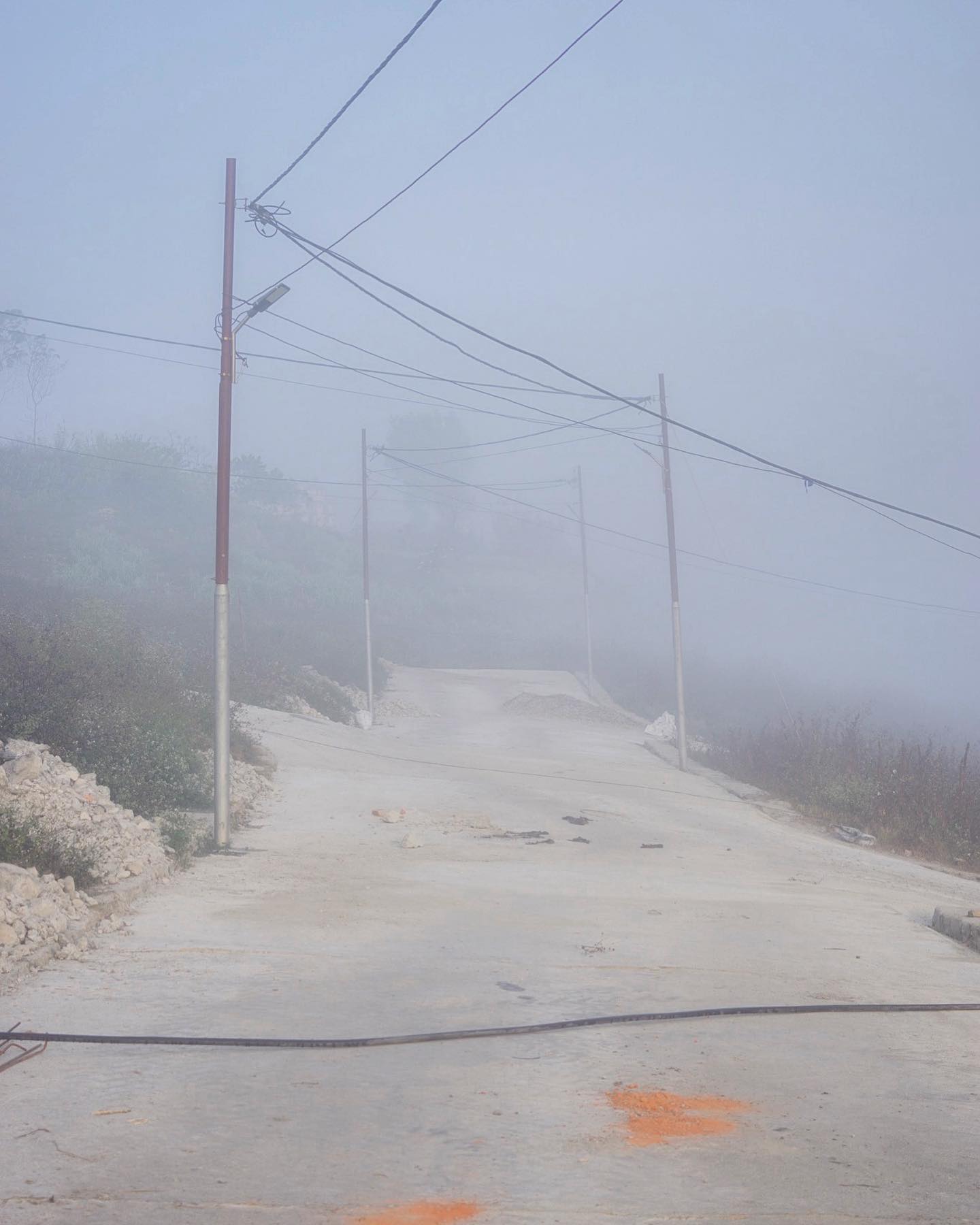 A concrete road leads uphill through dense fog with utility poles and power lines, creating a serene and mysterious scene.