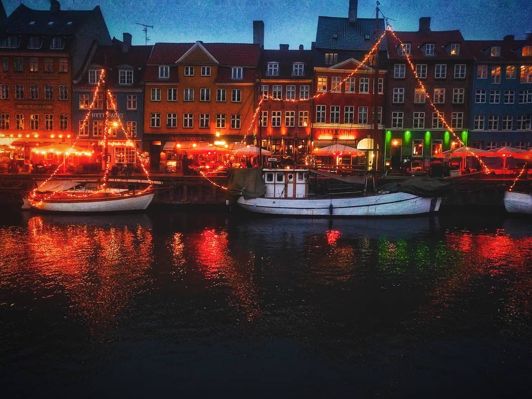 Boats docked in a canal in Copenhagen, Denmark, with colorful buildings lit up at night reflecting in the water.