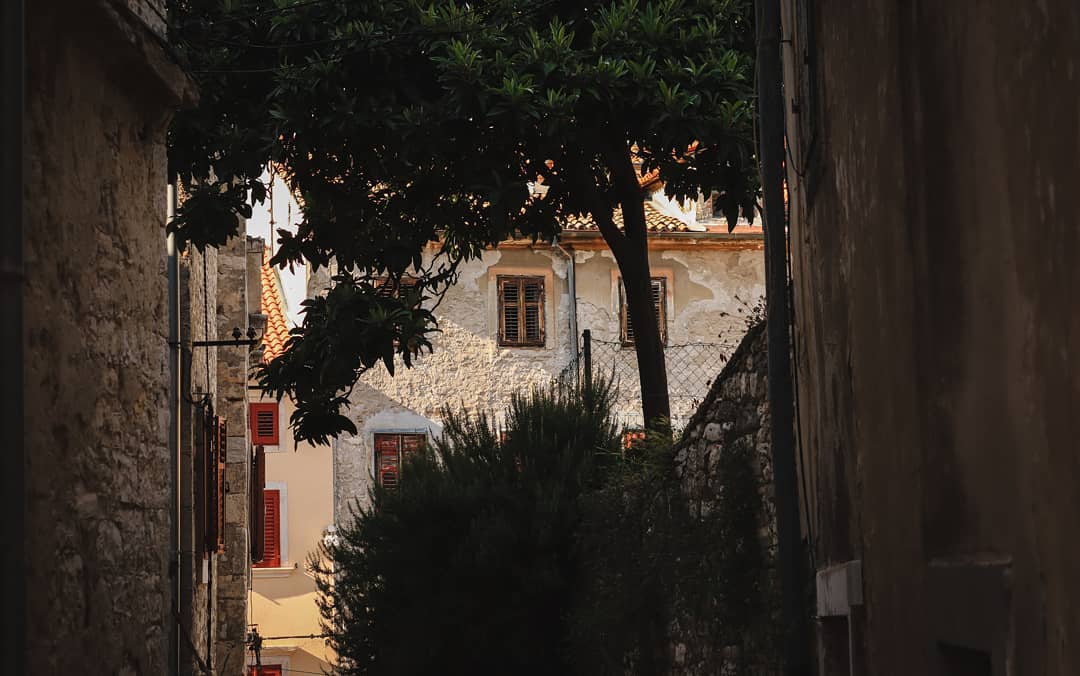 An alleyway view captures old buildings framed by greenery on a sunny day, promoting a sense of travel and heritage.