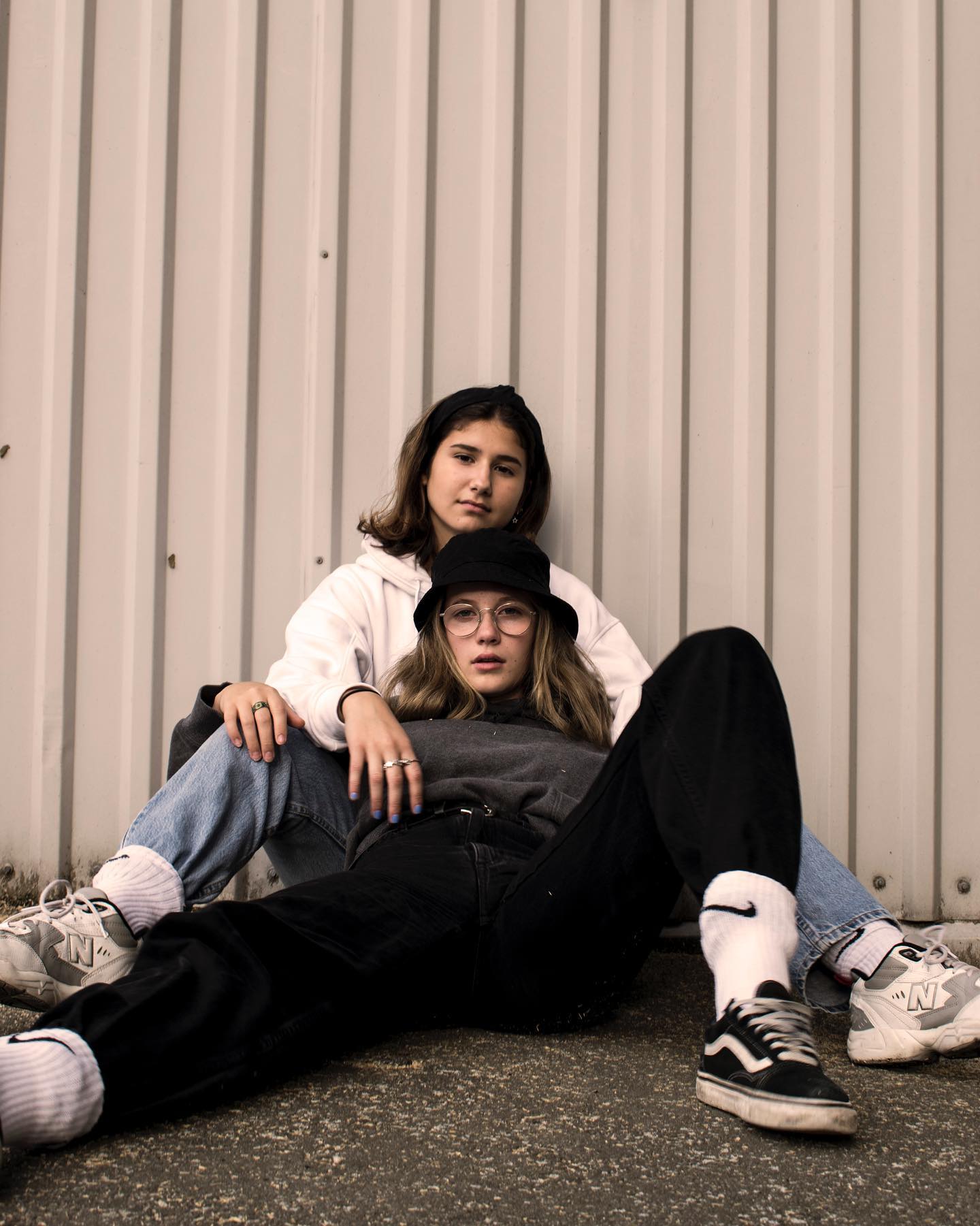 Two young women pose casually in front of a textured wall, showcasing a blend of urban street style.