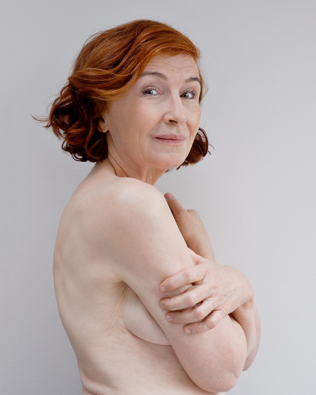 A redhead mature woman poses nude in a studio against a neutral background, radiating confidence and natural beauty.