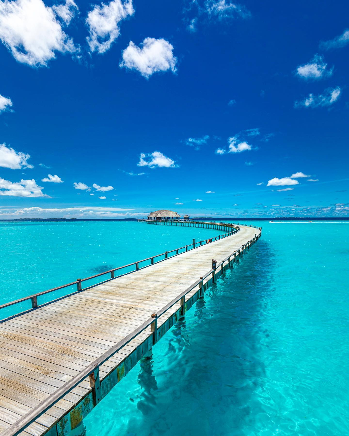 A beautiful wooden pier stretches over turquoise waters to a tropical island under a bright blue sky with fluffy clouds.