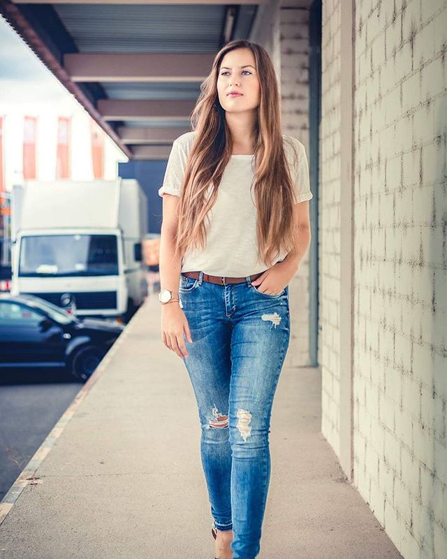 A young woman walks down a sidewalk wearing jeans and a t-shirt in an urban setting.