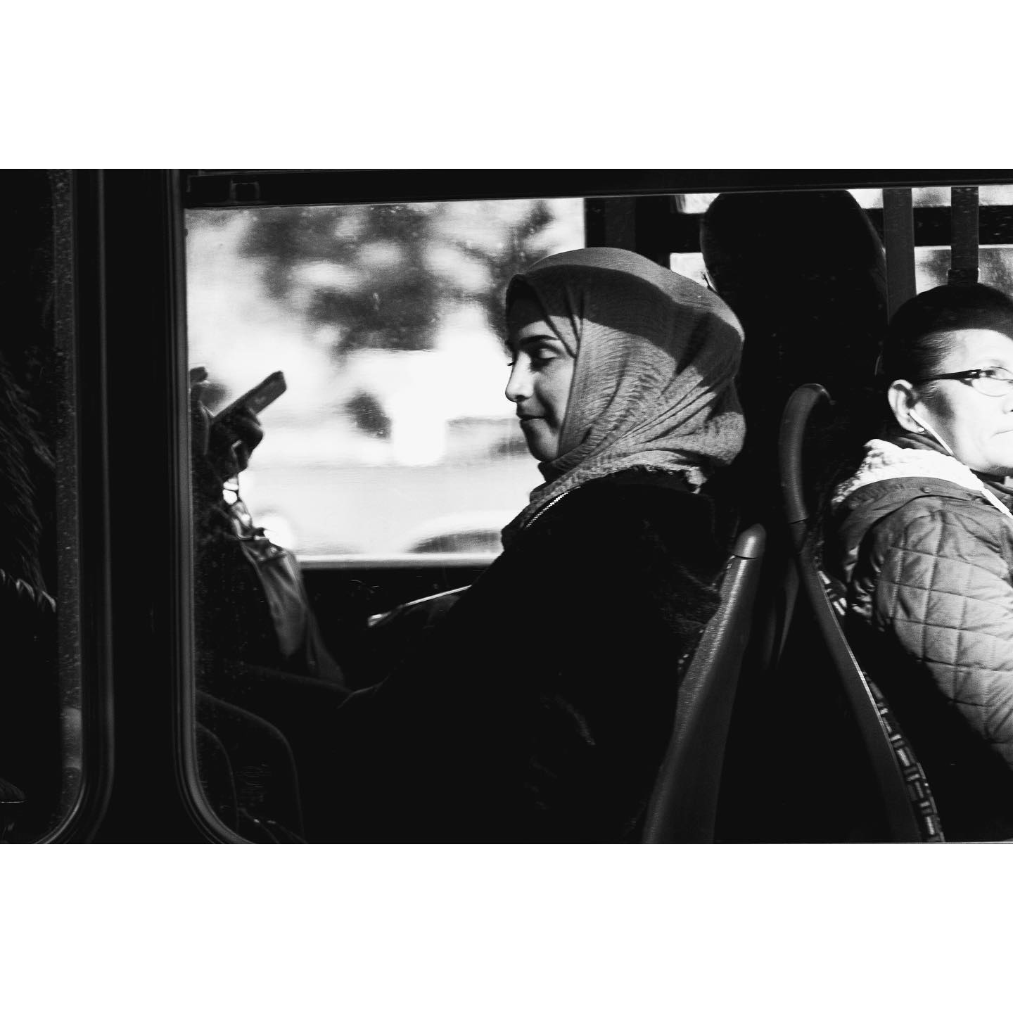 A woman wearing a hijab rides a bus, looking out the window in a black and white street photography style image.