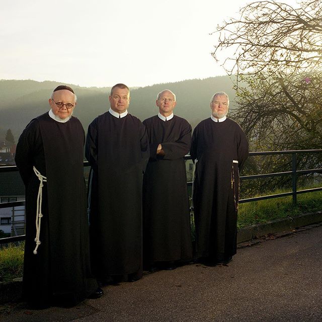 Four friars in black robes stand on a road against a rural backdrop. They are serious and look at the camera.