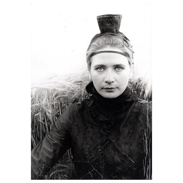 A woman with a unique headpiece poses in a field, captured in a timeless black and white portrait.