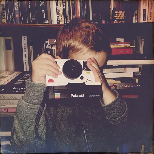 A young boy playfully takes a photo with a vintage Polaroid camera in front of a bookshelf.