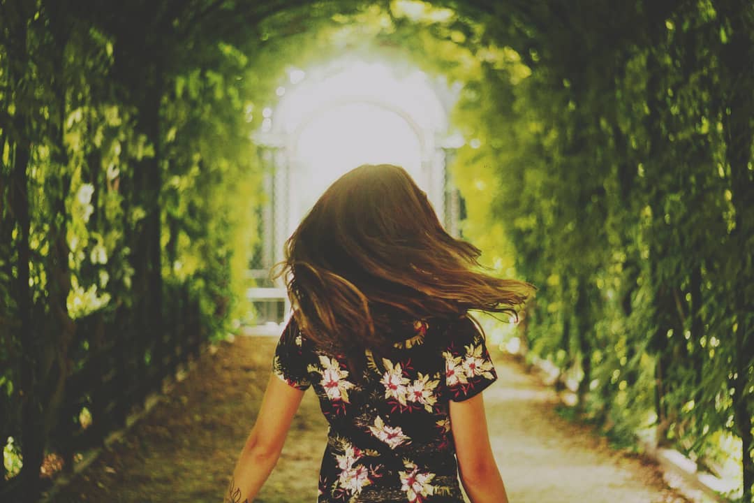 A woman walks through a lush green tunnel with sunlight streaming through, creating a sense of escape and tranquility.