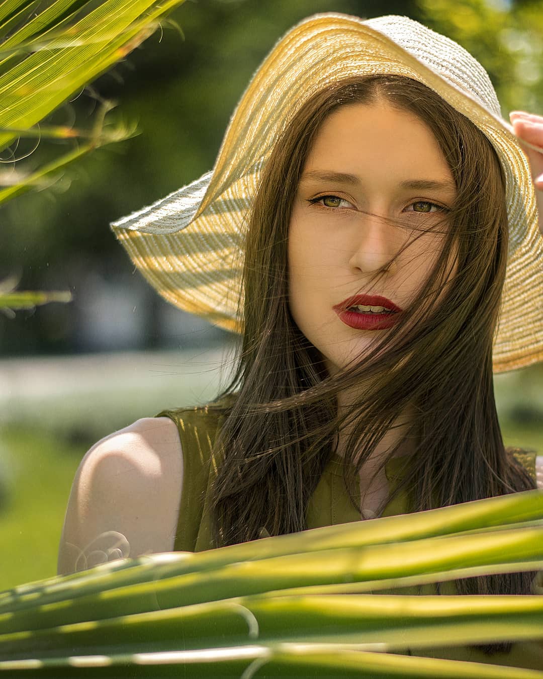 A woman wearing a hat with red lipstick poses outdoors surrounded by foliage in a soft, dreamy portrait.