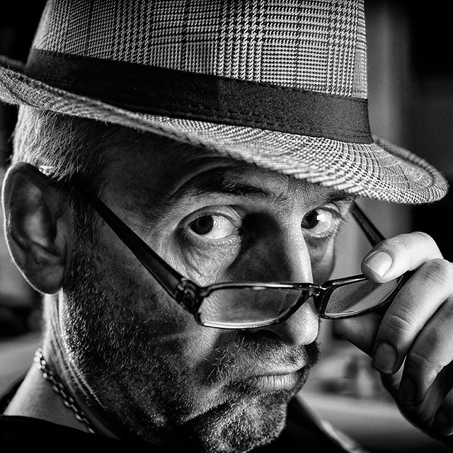 Intense black and white portrait of a man with a hat and glasses, looking directly at the camera.