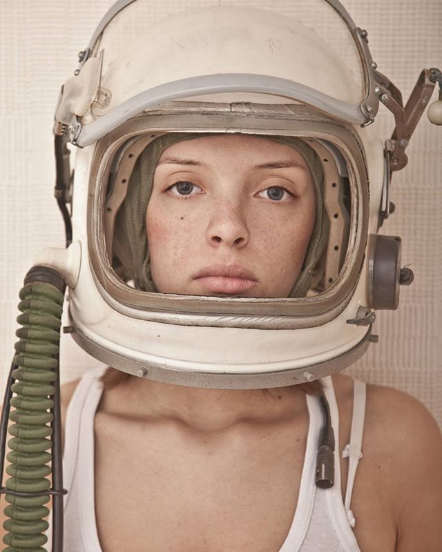 A woman wearing an old astronaut helmet with a white tank top. Shot with soft lighting and a neutral color palette.