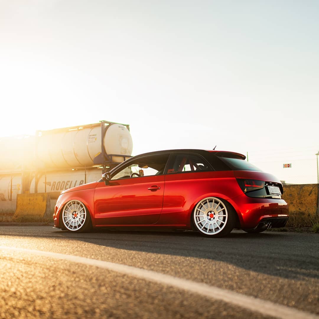 A stylish red Audi A1 hatchback sits on an asphalt road with modified wheels and suspension, bathed in warm sunlight.