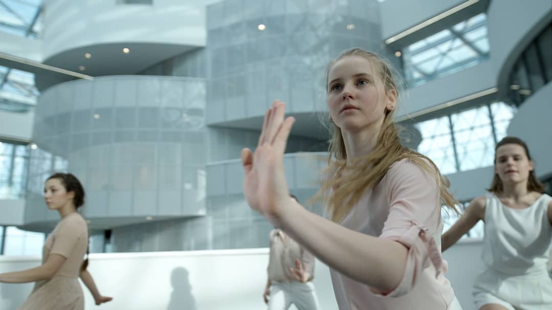 A group of young women gracefully dance in a modern, light-filled architectural space.