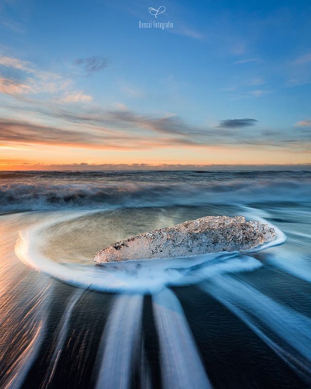 An ice block rests on a black sand beach as waves swirl around it in a long exposure shot.