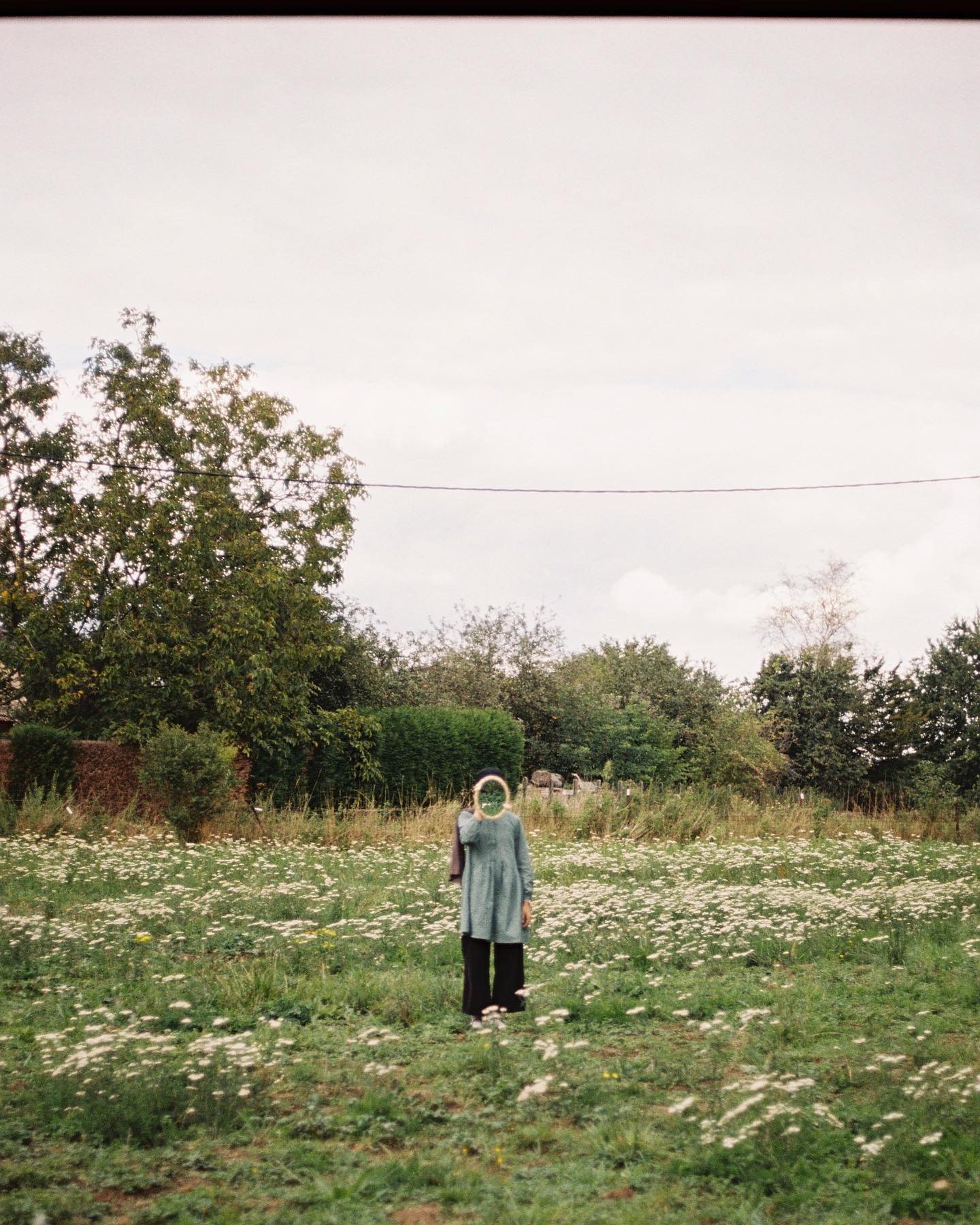 A woman in a field holds a mirror to her face, reflecting the natural surroundings in a dreamy, surreal scene.