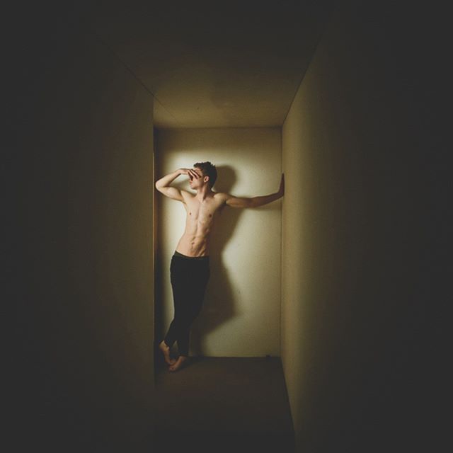 A shirtless man in black pants poses against a wall in a dimly lit room.