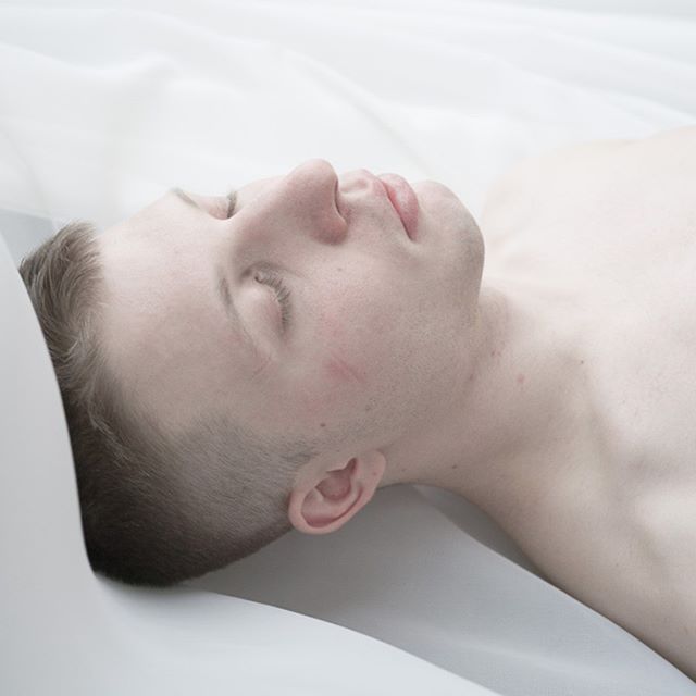 A man lying on a white sheet appears calm and serene, perfect for wellness ads.