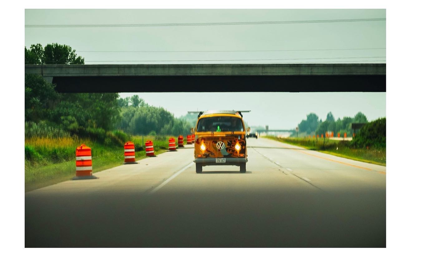 A vintage yellow Volkswagen bus drives down a highway lined with orange cones under a concrete underpass.