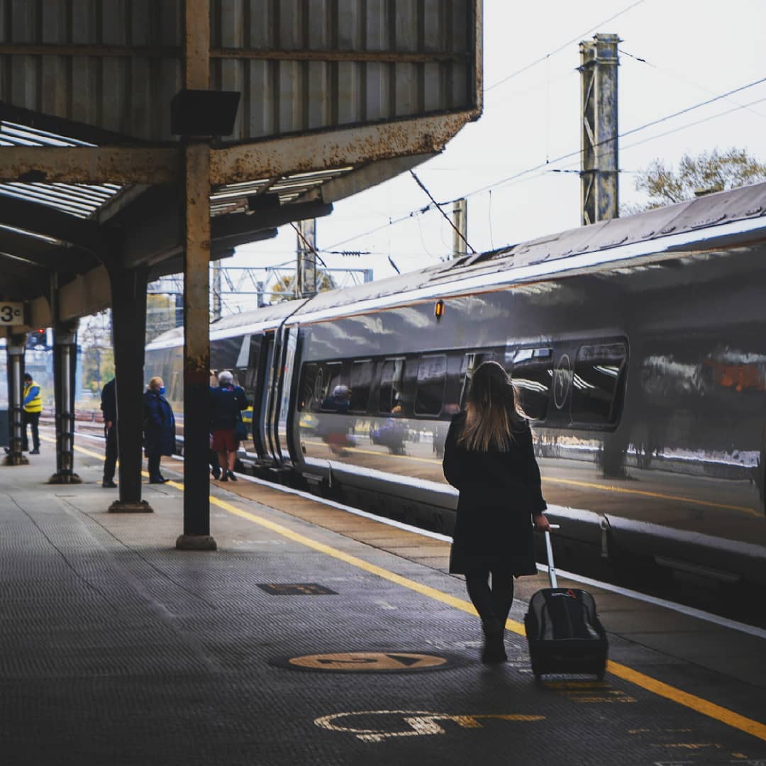 A woman walks with a suitcase along a train platform next to a modern train on a cloudy day.