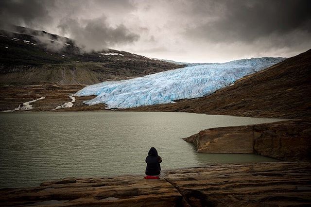 A lone figure sits by a glacial lake, surrounded by mountains and a glacier under a cloudy sky.