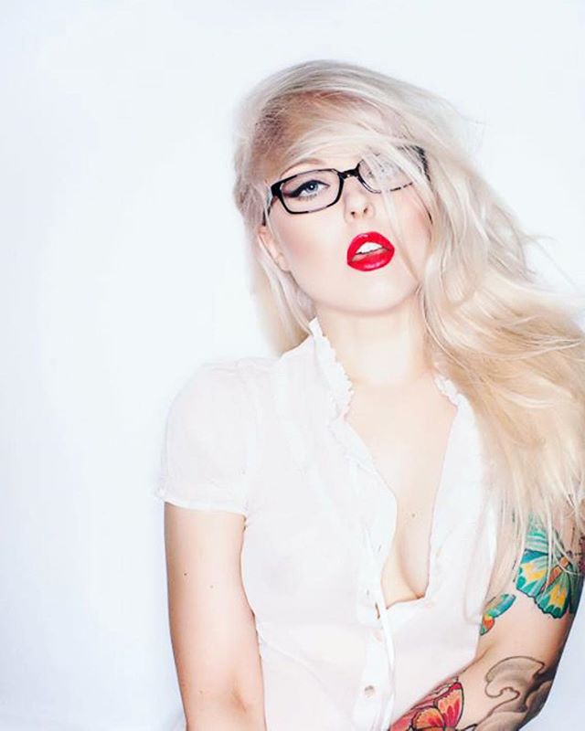 A blonde woman with red lipstick and glasses poses in a studio, showcasing a fashionable and confident style.
