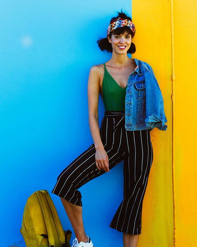 A smiling woman in a green top and striped pants poses against a colorful blue and yellow wall.