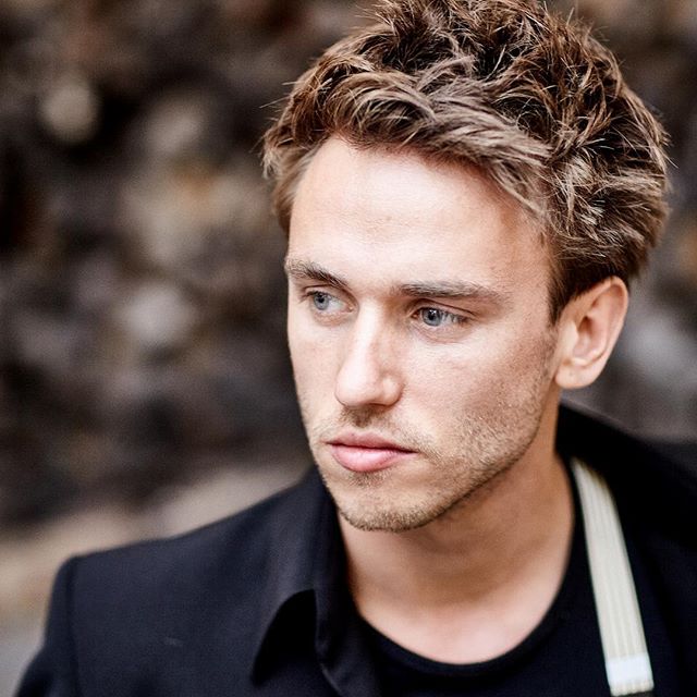 A serious-looking man with light hair and blue eyes in a portrait shot looking off to the side.