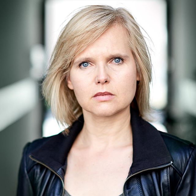 A serious woman with blonde hair poses for a professional headshot in a studio setting.