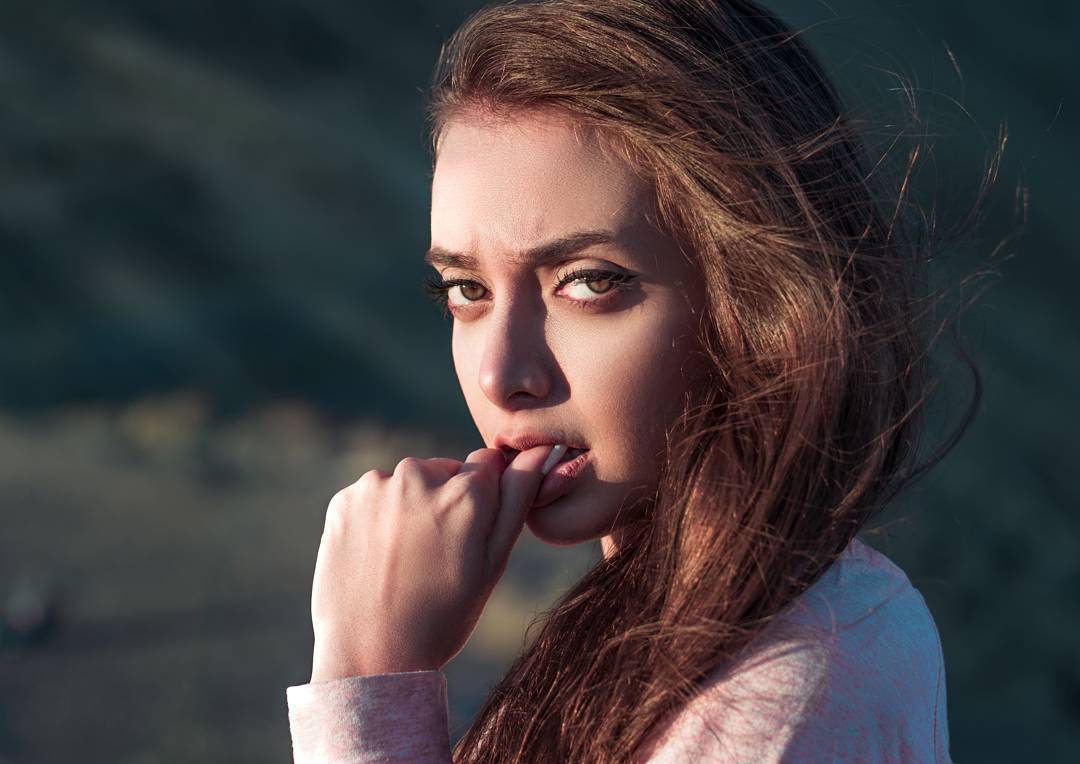 A brunette woman with an intense gaze and hand near her mouth poses outdoors in soft light.